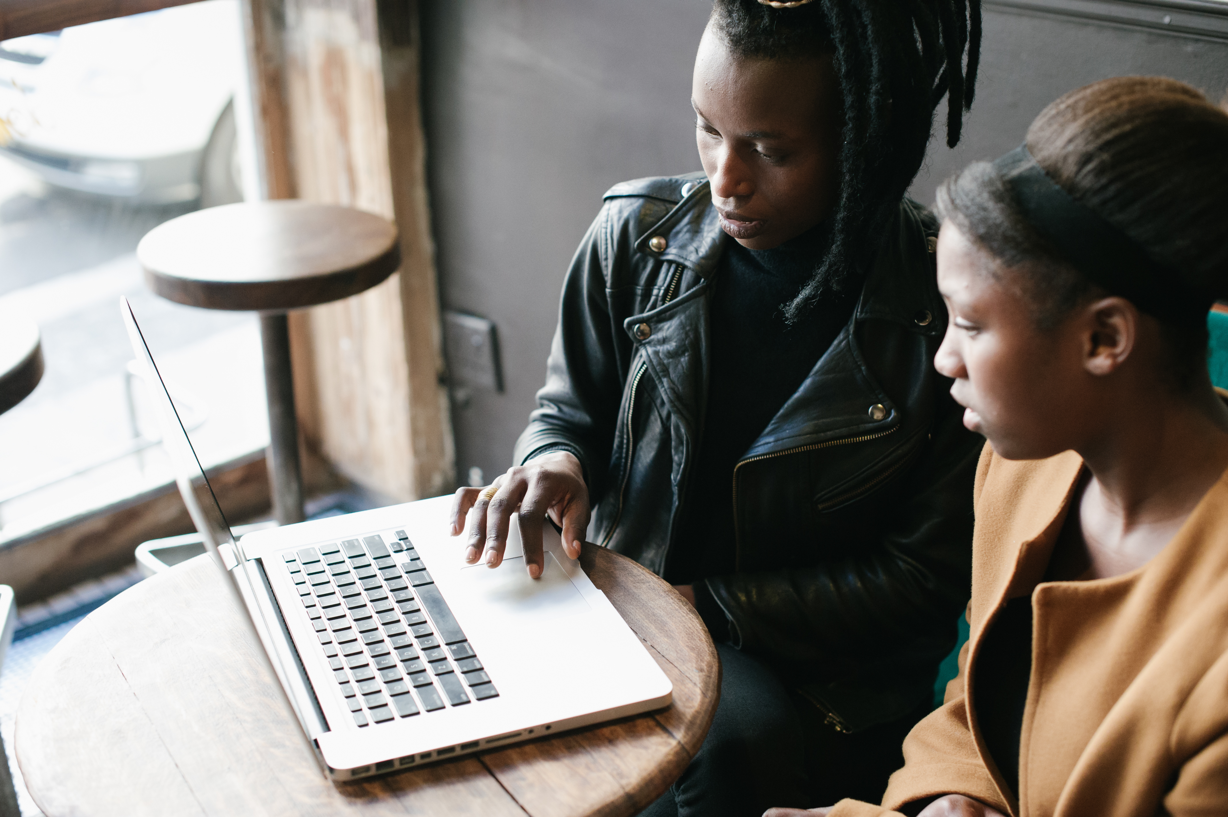 Two young black women sit at a round table, both reading from the same laptop. One, who is wearing a leather jacket, has her hand on the laptop trackpad.