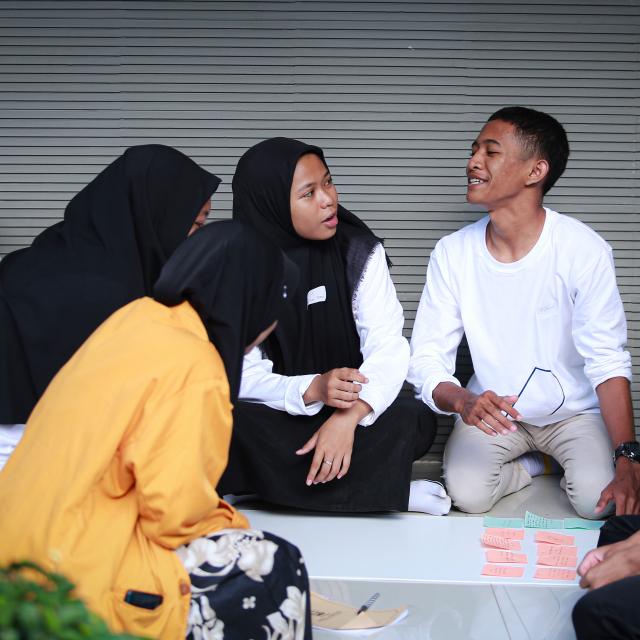 A group of people in an animated discussion, three are women in hijab, and all are kneeling infront of a poster board with post its lined up along the top.