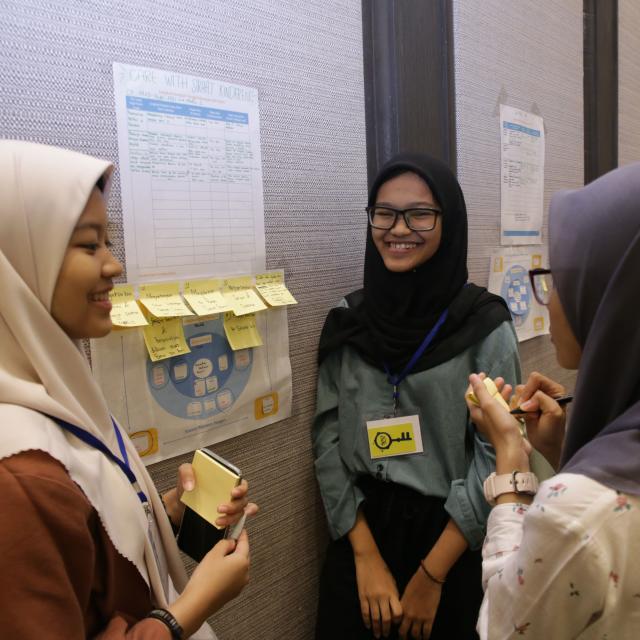 A group of women, all wearing hijab, standing at a poster with a diagram. They have placed several yellow sticky notes with text written on them on the poster. One woman is writing a new note on a sticky note pad.