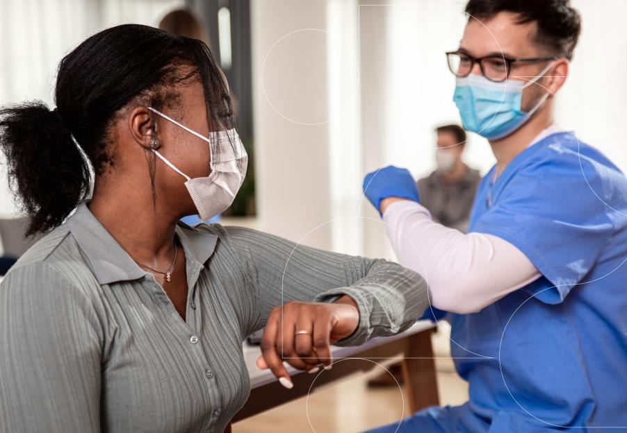 A medical worker and a patient, both wearing medical masks and sitting at a table, share an elbow bump greeting. 
