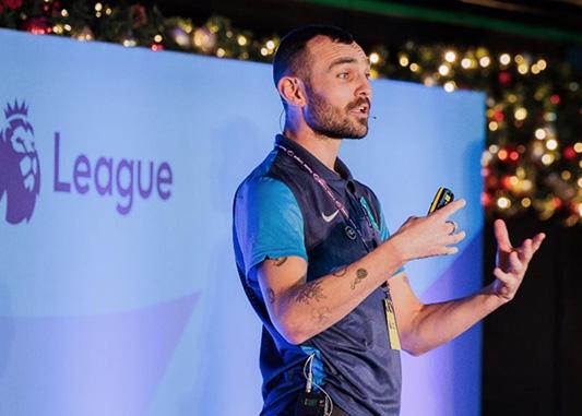A white man stands in front of a banner that with a portion of the Premier Leagues logo in frame. He is wearing a football jersey and jeans, and wears a headset and mic pack on his hip. He is speaking energetically, his hands are lifted in movement as he speaks to an unseen audience.