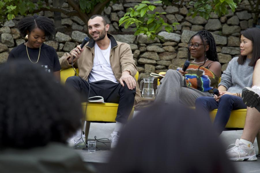 A group of young people of different races sit in low lounge chairs in an outdoor conversation space. In the foreground are the back of two people's heads, out of focus. One white man is holding a microphone, seemingly the speaker or giving a response.
