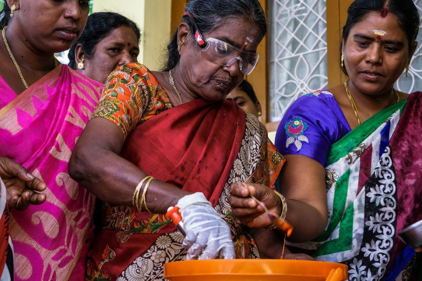 The image of several South Asian women in colorful sarongs. The woman at the center is wearing a latex glove and is mixing something in a large orange bucket.
