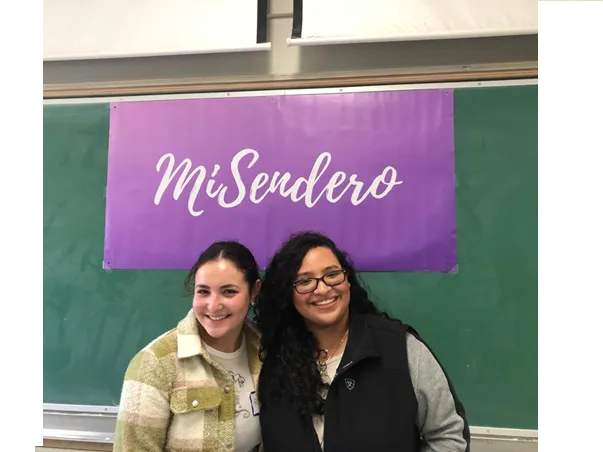 Romy Greenwald (left) and Jaqueline Gonzalez (right) at the 2022 El Congreso de UCSB Latine College Day. They are in front of a chalkboard with a purple poster that says "MiSendero." 