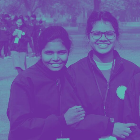 A stylized photo of two young Indian women wearing windbreakers, smiling and posing casually for a photo. In the distance behind the two girls is another group of young people wearing similar windbreaker jackets. The photo has been posterized in purple and teal, the Ambition Accelerator brand colors.
